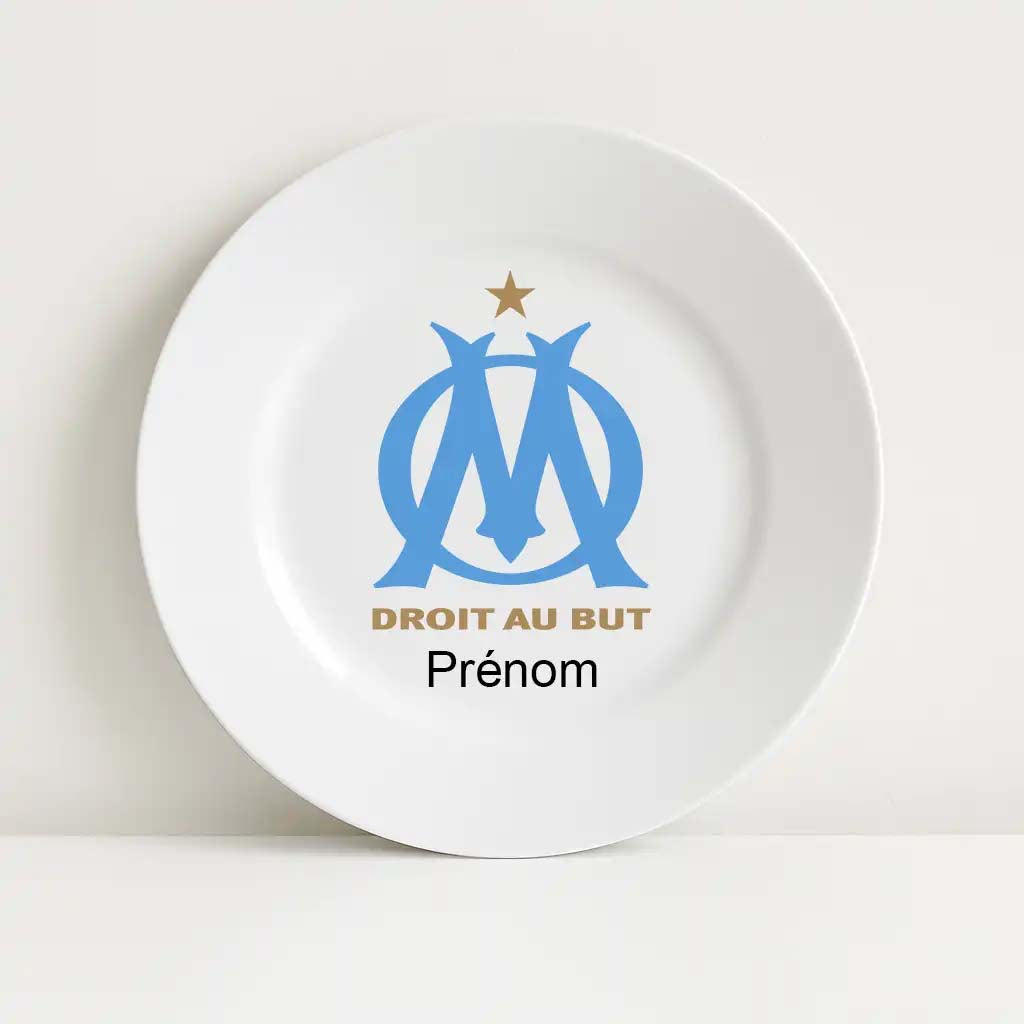 Assiette blanche incassable ornée du logo de l'Olympique de Marseille au centre. Le blason bleu ciel est accompagné de l'étoile dorée en haut et de la devise "DROIT AU BUT" en lettres dorées. En dessous, l’inscription "Prénom" est prévue pour être personnalisée.